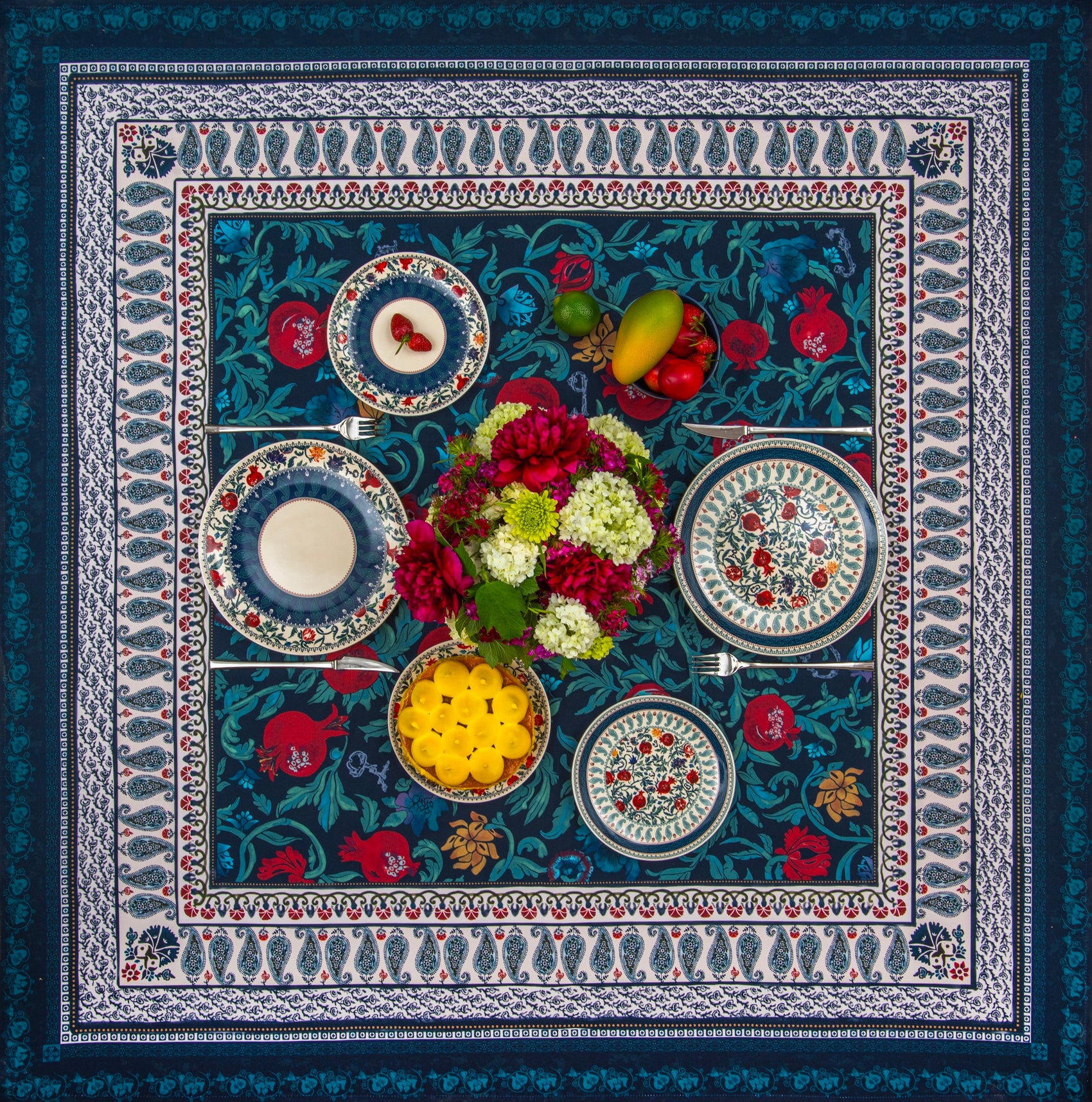 Flat lay of the BlueTabla blue pomegranate tablecloth, set with four patterned plates, a floral centrepiece, fruit and desserts.