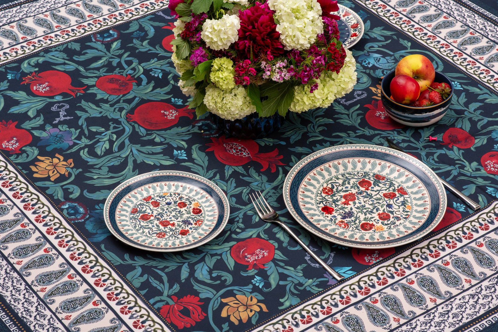 BlueTabla Georgian Alphabet & Pomegranate blue tablecloth styled with stacked patterned plates, apples and a peony bouquet.