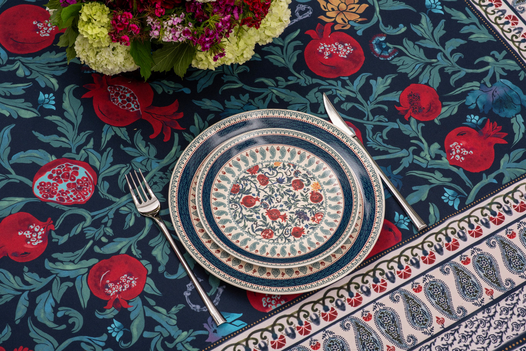 Place setting on the BlueTabla blue pomegranate tablecloth with cutlery, a peony bouquet and the paisley border in view.