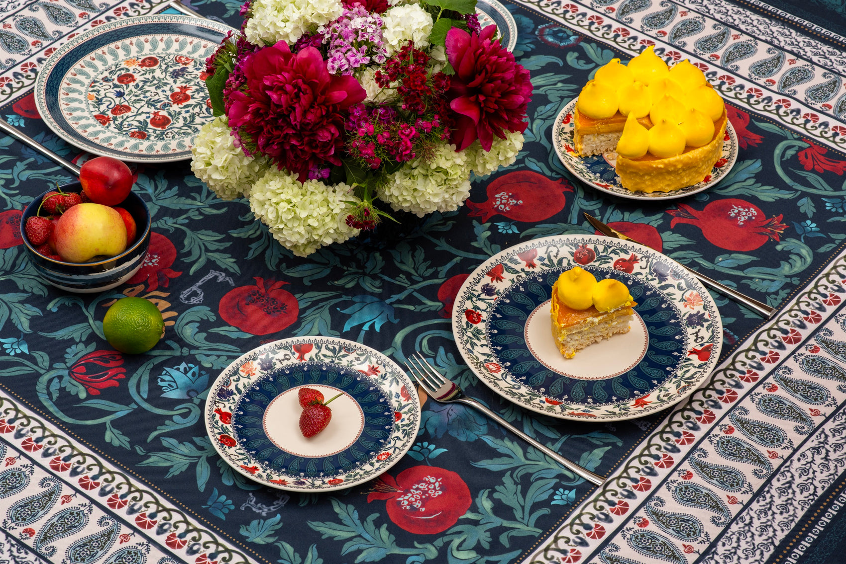 BlueTabla blue tablecloth set for tea with patterned plates, a peony and hydrangea bouquet, fresh fruit and lemon desserts.
